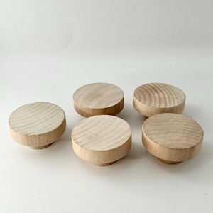 May include: Five unfinished wooden knobs, each with a round top and a slightly raised base. The knobs are arranged in a semi-circle on a white surface.