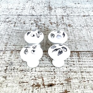 Peut inclure: Quatre boutons en c&eacute;ramique blanche avec des motifs de coquillages noirs et gris. Les boutons sont dispos&eacute;s en deux paires, les boutons du haut &eacute;tant orient&eacute;s vers l'avant et les boutons du bas &eacute;tant orient&eacute;s vers l'arri&egrave;re.