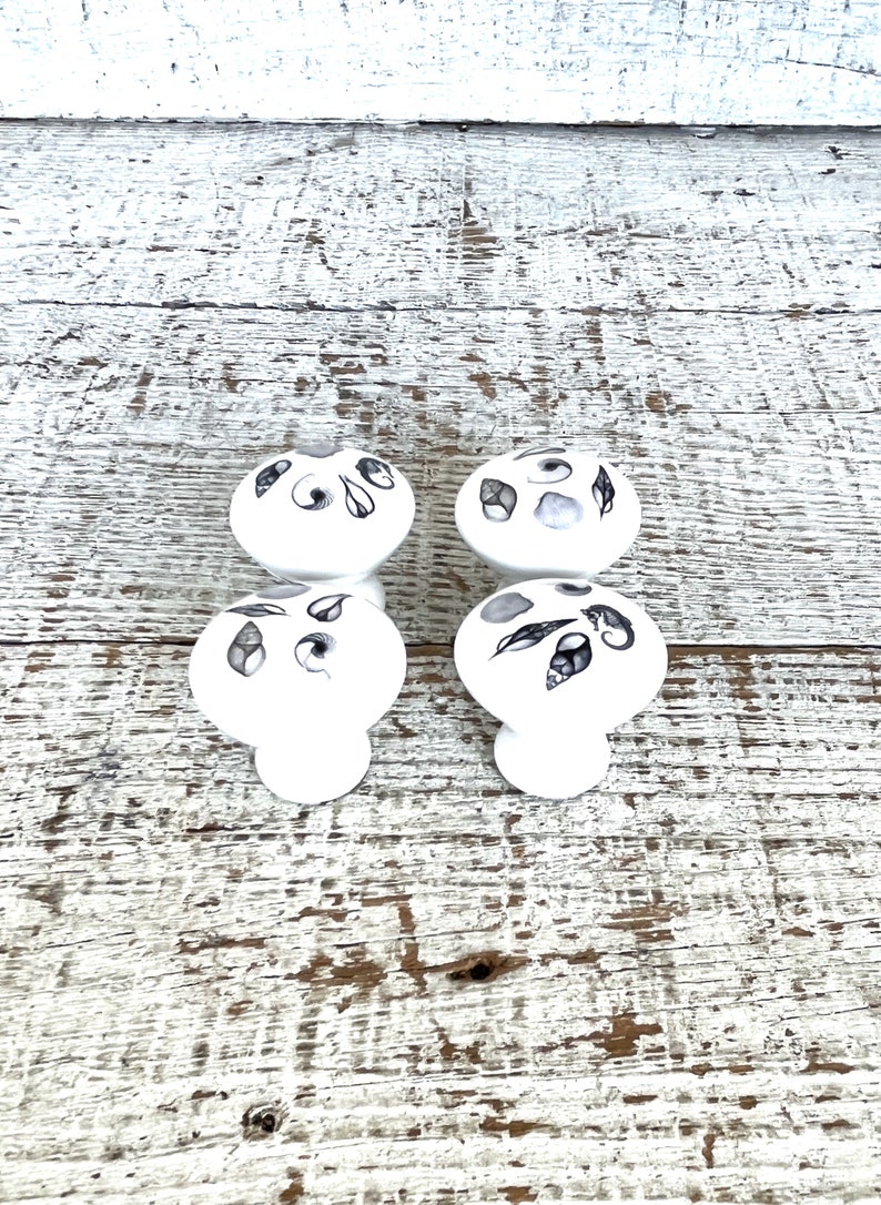 Peut inclure: Quatre boutons en c&eacute;ramique blanche avec des motifs de coquillages noirs et gris. Les boutons sont dispos&eacute;s en deux paires, les boutons du haut &eacute;tant orient&eacute;s vers l'avant et les boutons du bas &eacute;tant orient&eacute;s vers l'arri&egrave;re.