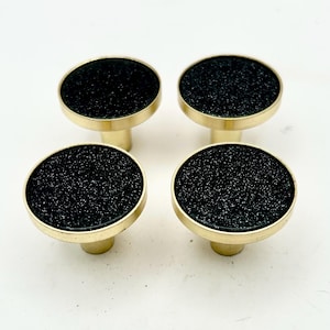 May include: Set of four cabinet knobs. Each knob features a round, black, textured surface set within a gold-colored metal frame. The knobs have a cylindrical base, and are suitable for furniture or cabinet hardware.