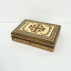 May include: A rectangular wooden box with a decorative inlaid design. The lid features a light-colored cross design within a darker border. The box is made of various wood tones and has intricate patterns.