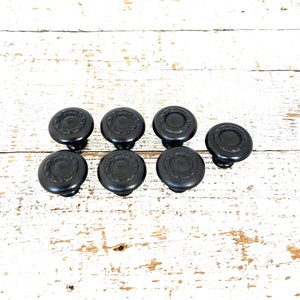 May include: Eight black cabinet knobs with a decorative raised design. The knobs are arranged in two rows of four on a white wooden surface.