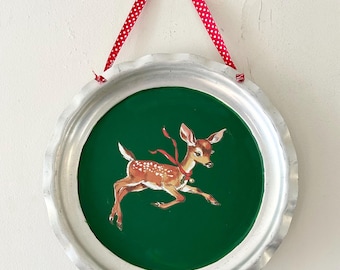 Puede incluir: Un adorno de pared redondo de metal plateado con un centro verde que presenta una ilustración de un cervatillo saltando. El cervatillo tiene manchas marrones y una cinta roja. Se utiliza un lazo rojo y una cinta de lunares para colgarlo.