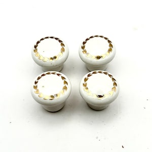 May include: Four white ceramic cabinet knobs with a gold laurel wreath design. The knobs are round and have a classic, decorative style. Ideal for adding a touch of elegance to furniture or cabinetry.