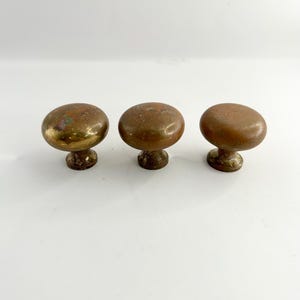 May include: Three antique brass cabinet knobs. Each knob has a rounded top and a short, decorative base. The knobs are arranged in a row against a white background. The brass has a warm, aged patina.