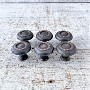 May include: Six black cabinet knobs with a copper colored ring around the edge. The knobs have a slightly raised design on the top.