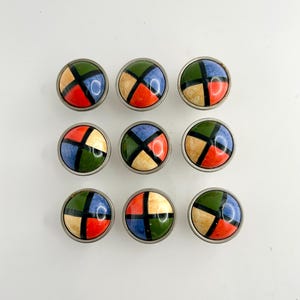 Puede incluir: Nueve pomos de armario redondos con un diseño geométrico colorido. Cada pomo presenta una base de metal plateado y una parte superior abovedada brillante dividida en cuadrantes de rojo, azul, verde y beige, separados por líneas negras.