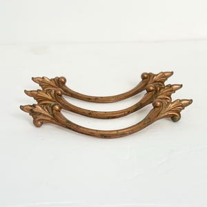 May include: Three curved drawer pulls made of aged bronze-toned metal. Each handle features a decorative leaf-like motif at both ends with a smooth, arched center bar. The metal surface shows signs of wear and patina, giving it a weathered appearance.