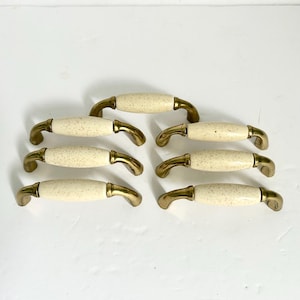 May include: A set of six cabinet pulls featuring a central cylindrical cream-colored ceramic handle with brown speckles, flanked by curved brass-toned metal ends. Each handle is mounted on two metal posts, creating a uniform arch shape.
