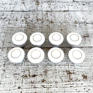 May include: Eight white ceramic knobs with a gold trim. The knobs are arranged in two rows of four on a whitewashed wooden surface.