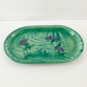 Könnte beinhalten: Ovale grüne Keramikschale mit handgemaltem Design aus lila Disteln und farnartigem Laub. Die Schale hat eine glänzende Oberfläche und einen leicht erhöhten Rand. Der Hintergrund ist ein hellerer Grünton.