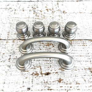 May include: A collection of six brushed nickel cabinet hardware pieces. The set includes four round knobs and two curved pull handles. The hardware is displayed on a distressed white wooden surface.