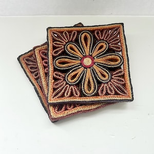Könnte beinhalten: Drei quadratische Untersetzer mit floralem Design. Sie bestehen aus gewebten Fasern in Braun-, Schwarz- und Rottönen. Die Mitte jedes Untersetzers zeigt eine Blume mit roter Mitte und Blütenblättern in Braun- und Schwarztönen.