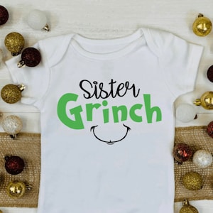 May include: A white baby bodysuit with the words "Sister Grinch" in black and green, and a smiling face graphic. The image is decorated with gold, white, and red Christmas baubles.