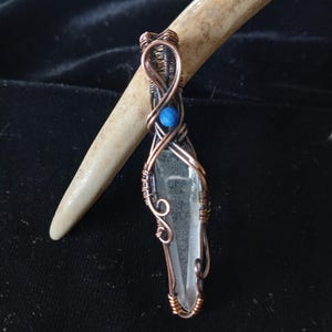May include: A clear quartz crystal pendant wrapped in copper wire. The wirework features intricate swirls and a small blue bead. The pendant is suspended from a copper wire loop. The background is a dark, textured fabric.