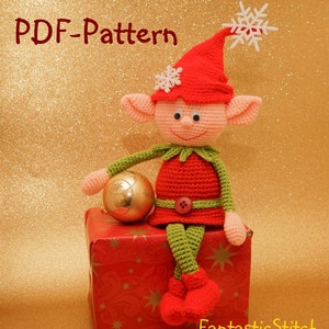 Könnte beinhalten: Häkelanleitung für eine Weihnachtswichtelpuppe mit roter Mütze, grüner Hose und einem rot-grünen Outfit. Der Wichtel hält einen goldenen Anhänger und sitzt auf einer roten Geschenkbox vor goldenem Hintergrund.