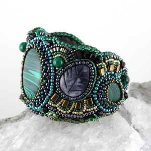 May include: A beaded bracelet with green, black, and gold accents. The bracelet features a large, oval-shaped malachite stone and a smaller, round malachite stone. The bracelet is also decorated with small, black beads and gold beads.