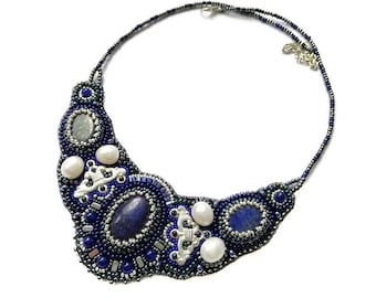 Puede incluir: Un collar de cuentas con una piedra azul grande y detalles plateados. El collar está hecho con cuentas azul oscuro, azul claro y plateadas. El collar es una pieza llamativa y sería perfecto para una ocasión especial.