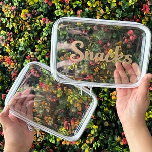 May include: Two clear rectangular pouches with white trim and zippers. The word "Snacks" is written in a gold script across the front of one pouch. The pouches are held up against a background of colorful foliage.