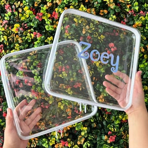 May include: Clear, square-shaped cosmetic bags with white trim and zippers. One bag has the name "Zoey" in blue script. The bags are held up against a backdrop of colorful foliage.