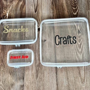 May include: Three clear, rectangular zippered pouches with white trim. The largest pouch has the word "Crafts" in black. The medium pouch has "Snacks" in gold, and the smallest pouch has "FIRST AID" in red.