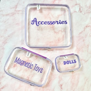 May include: Three clear, rectangular storage cases with purple trim and silver clasps. The largest case has the word "Accessories" in purple script. The second case says "Magnetic Toys", and the smallest says "DOLLS".