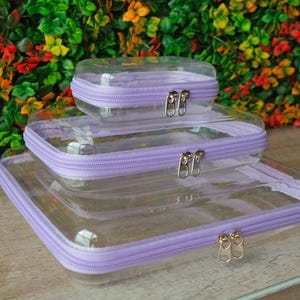 May include: Three clear plastic zippered pouches with lavender trim. The pouches are nested inside each other, with the largest pouch on the bottom and the smallest pouch on top.