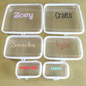 May include: Six clear plastic zippered pouches with white trim. The pouches are labeled with the words "Zoey", "Crafts", "Snacks", "Kylie", "First Aid", and "Cards".