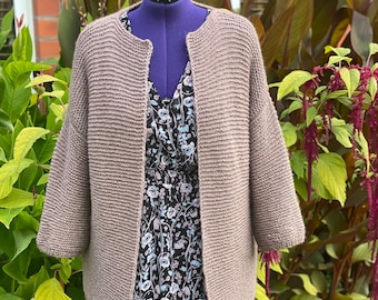 May include: A brown knitted cardigan with a slightly open front and long sleeves. The cardigan is displayed on a mannequin against a backdrop of green foliage.