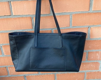 May include: A black leather tote bag with a single pocket on the front. The bag is hanging from a hook on a brick wall.