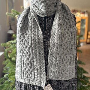 May include: A gray cable-knit scarf with a matching cowl. The scarf features a detailed woven pattern and is displayed on a dress form. A small tag hangs from the scarf. The background includes a Christmas tree.