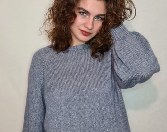 Puede incluir: Una mujer con un suéter gris con un brillo sutil y pantalones vaqueros negros. El suéter tiene un cuello redondo y mangas largas.