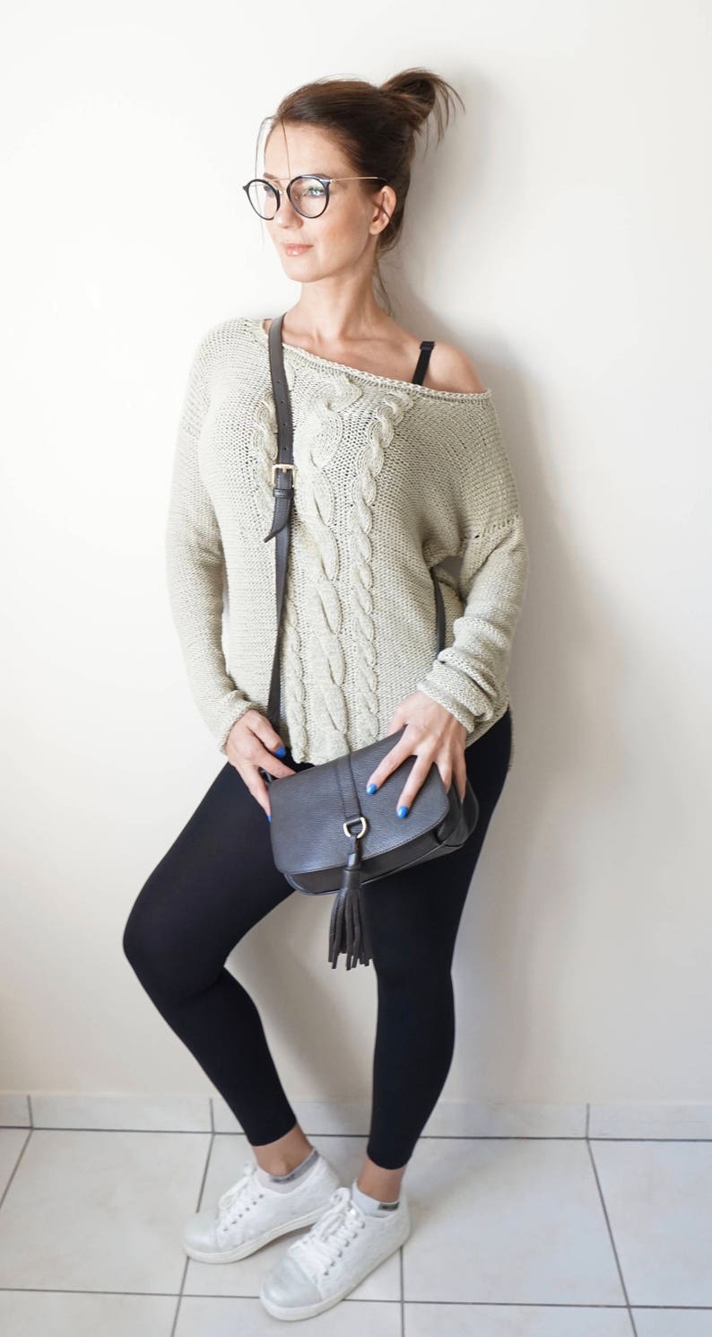 May include: A woman wearing a light beige cable knit sweater with a black leather shoulder bag, black leggings, and white sneakers.