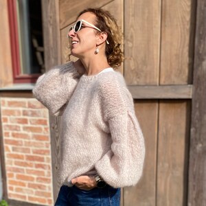 Puede incluir: Una mujer lleva un su&eacute;ter de color rosa claro y peludo con mangas largas y cuello redondo. Lleva gafas de sol blancas y pendientes de perlas. Est&aacute; de pie frente a una pared de madera.