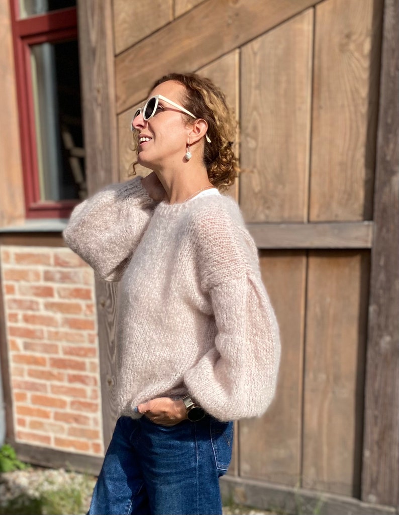 Puede incluir: Una mujer lleva un su&eacute;ter de color rosa claro y peludo con mangas largas y cuello redondo. Lleva gafas de sol blancas y pendientes de perlas. Est&aacute; de pie frente a una pared de madera.