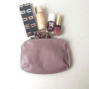 May include: A collection of beauty products including a foundation bottle, two nail polish bottles, a Benefit lip stain, and a Bobbi Brown eyeshadow palette. A mauve-colored zippered pouch and a patterned scrunchie are also present.