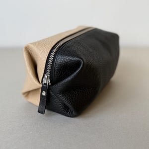 May include: A two-tone leather pouch, half beige and half black, with a silver zipper closure. The pouch is rectangular with a rounded shape and a small leather pull tab on the zipper.