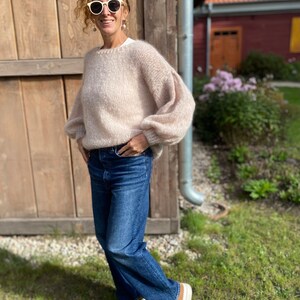 Puede incluir: Una mujer con un su&eacute;ter de punto rosa claro con mangas abullonadas, jeans azules y zapatillas marrones y blancas. Est&aacute; de pie frente a una pared de madera y lleva gafas de sol blancas.