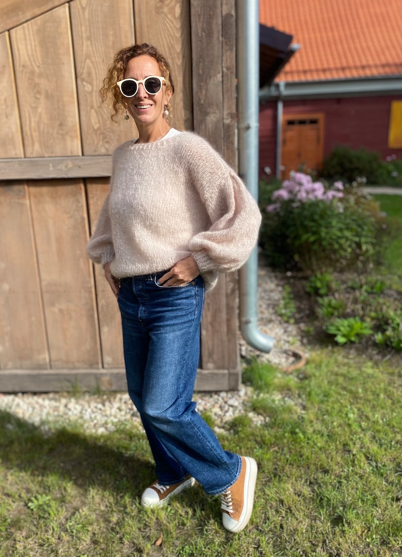 Puede incluir: Una mujer con un su&eacute;ter de punto rosa claro con mangas abullonadas, jeans azules y zapatillas marrones y blancas. Est&aacute; de pie frente a una pared de madera y lleva gafas de sol blancas.