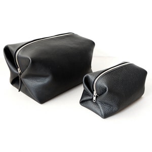 May include: Two black leather cosmetic bags with silver zippers. The larger bag is rectangular, and the smaller bag is square. Both bags have a textured surface and are set against a white background.