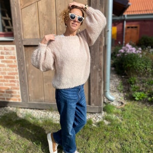 Puede incluir: Una mujer lleva un su&eacute;ter de punto rosa claro con mangas grandes, jeans azules y zapatillas blancas. Est&aacute; de pie frente a un edificio de madera con una pared de ladrillo.