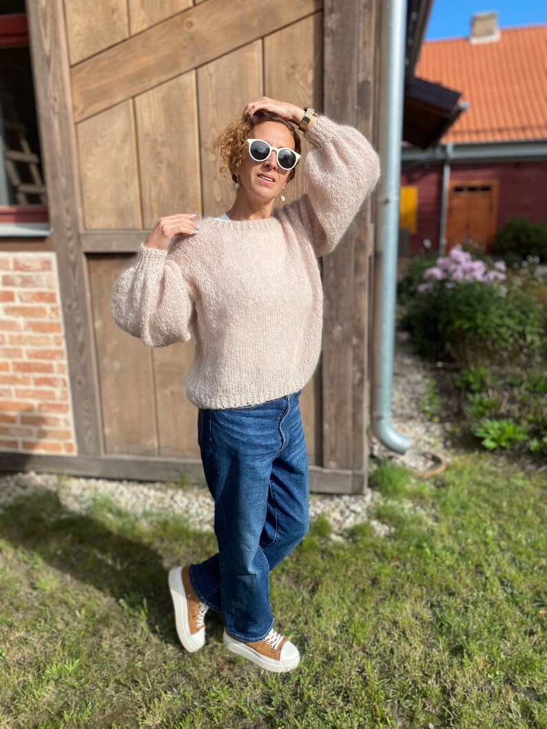 Puede incluir: Una mujer lleva un su&eacute;ter de punto rosa claro con mangas grandes, jeans azules y zapatillas blancas. Est&aacute; de pie frente a un edificio de madera con una pared de ladrillo.