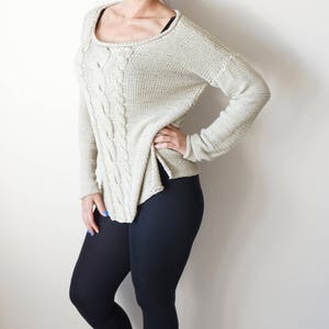 May include: A woman wearing a light beige cable knit sweater with black leggings and white sneakers.