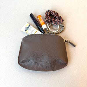 May include: A brown leather cosmetic bag with a zipper, containing beauty products and accessories. Items include a tube of cream, mascara, lip balm, a scrunchie, and beaded bracelets. The bag is on a neutral surface.