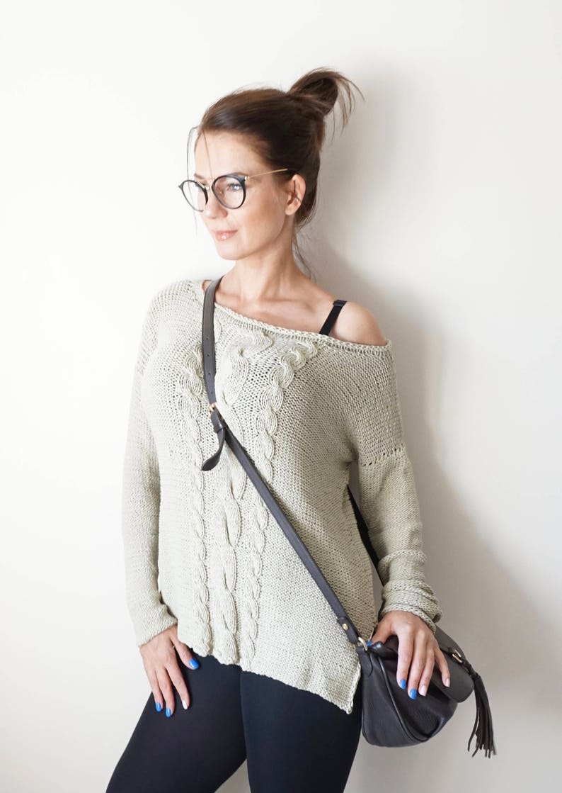 May include: A woman wearing a light green cable knit sweater with a black leather crossbody bag. She is wearing black leggings and has her hair pulled back in a bun. She is wearing black framed glasses.