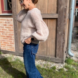 Puede incluir: Una mujer con un su&eacute;ter de color rosa claro y peludo con jeans azules y zapatillas marrones. Est&aacute; de pie frente a una pared de madera.