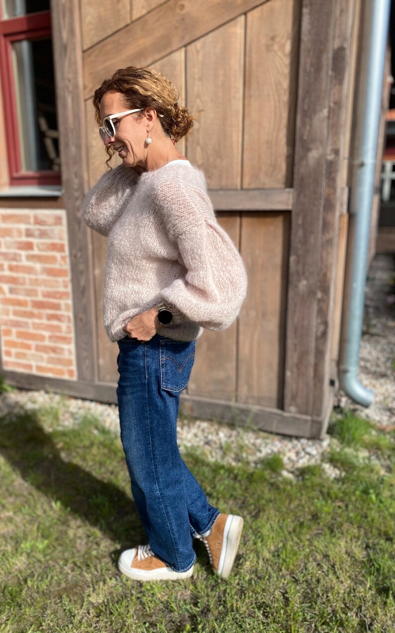 Puede incluir: Una mujer con un su&eacute;ter de color rosa claro y peludo con jeans azules y zapatillas marrones. Est&aacute; de pie frente a una pared de madera.