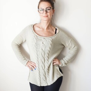 May include: A woman wearing a light beige cable knit sweater with black leggings and round glasses.