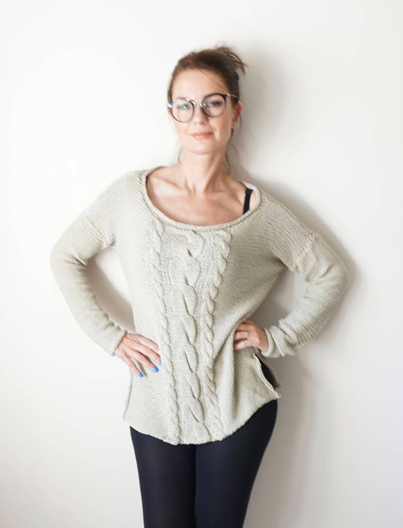 May include: A woman wearing a light beige cable knit sweater with black leggings and round glasses.