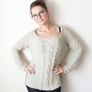 May include: A woman wearing a light beige cable knit sweater with long sleeves. She is wearing black pants and has her hair pulled back in a bun. She is wearing black framed glasses and has blue nail polish.